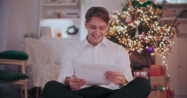 Boldog fiatalember olvas levelet, miközben ül díszített otthon karácsonykor - Felvétel, videó