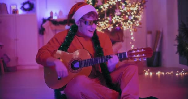 Glücklicher junger Mann mit Nikolausmütze und Sonnenbrille, der zu Weihnachten auf dem Fußboden im beleuchteten Haus sitzt und Gitarre spielt - Filmmaterial, Video
