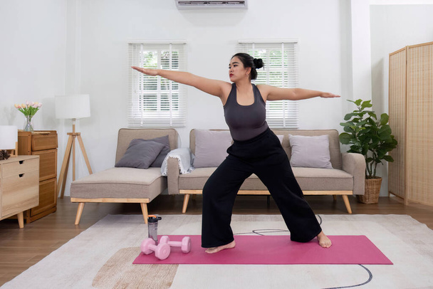 Dagadt ázsiai nő nyújtózkodik otthon egy fitneszszőnyegen. Gyakorló tevékenységek otthon online gyakorló órák Gyakorlás nyújtózkodik a jóga szőnyegen otthon, hogy egészséges és formás maradjon.. - Fotó, kép