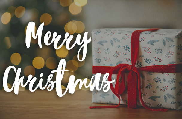 Καλά Χριστούγεννα κείμενο στο κομψό χριστουγεννιάτικο δώρο με κόκκινη κορδέλα κοντά ενάντια χριστουγεννιάτικο δέντρο φώτα bokeh. Καλές γιορτές! Ευχετήρια κάρτα εποχής. Χειρόγραφο σήμα - Φωτογραφία, εικόνα