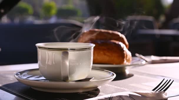 Stilleven. Langzame beweging. Witte kop warme drank, thee of koffie americano met stoom op een achtergrond van wazig Franse croissants, geserveerd als ontbijt in een gezellig café buiten. Voedsel- en drankconcept - Video