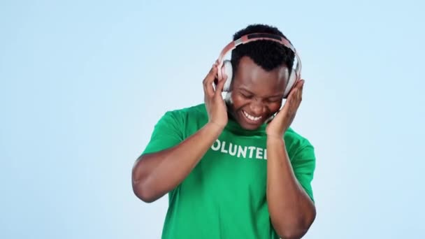 Música, danza y sonrisa con un hombre negro voluntario en un fondo azul en el estudio para la libertad o la caridad. Comunidad, energía y un joven feliz transmitiendo audio con auriculares para el bienestar. - Imágenes, Vídeo