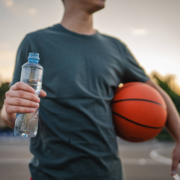 Kaukasier Mann Teenager öffnen Plastikflasche Wasser im Freien in sonnigen Tag trinken, während halten Basketball - Foto, Bild