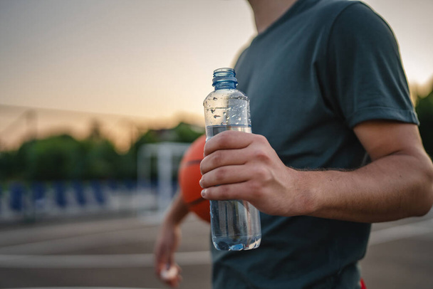caucásico hombre adolescente abierto botella de plástico de agua al aire libre en día soleado beber mientras mantenga baloncesto - Foto, imagen