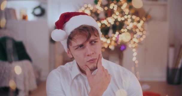 Στοχαστικός νεαρός με το χέρι στο πηγούνι φορώντας το καπέλο του Αϊ Βασίλη τα Χριστούγεννα στο σπίτι - Πλάνα, βίντεο