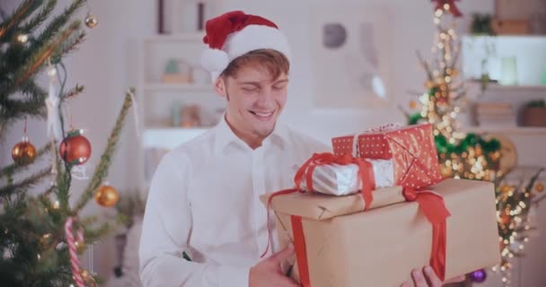 Ritratto di bel giovane in cappello di Babbo Natale sorridente mentre porta vari regali di Natale a casa  - Filmati, video