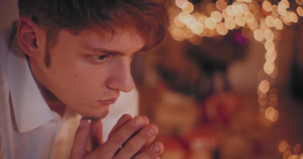 zbliżenie zamyślony młody człowiek z rękami zaciśnięty podczas Boże Narodzenie w domu - Materiał filmowy, wideo