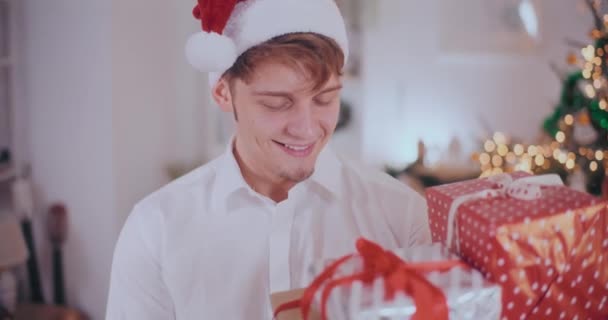 Retrato del joven guapo en el sombrero de Santa sonriendo mientras mira los regalos de Navidad en casa - Imágenes, Vídeo