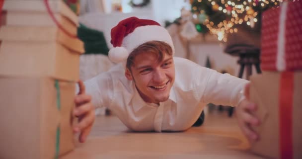 Πορτρέτο του κατάπληκτος νεαρός άνδρας σε Σάντα καπέλο συρόμενα δώρα Χριστουγέννων, ενώ βρίσκεται στο πάτωμα στο σπίτι - Πλάνα, βίντεο