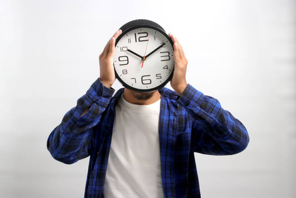 Азиат, одетый в шапочку и повседневную рубашку, креативно использует часы, чтобы прикрыть голову или лицо, держа его перед собой, стоя на белом фоне. Концепция управления временем - Фото, изображение