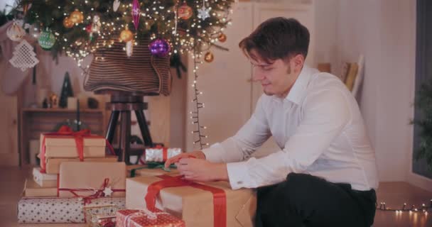 Feliz joven en formales atando cinta en caja de regalo envuelta mientras está sentado en el suelo en casa durante la Navidad - Imágenes, Vídeo