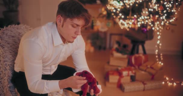 Joven deprimido sentado con la cabeza en las manos en la silla en casa con el árbol de Navidad iluminado en el fondo - Imágenes, Vídeo
