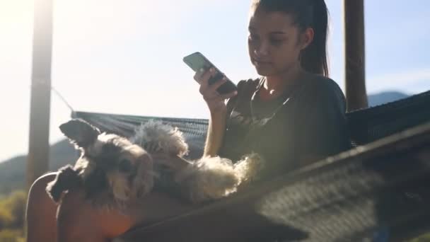 Woman, dog and relax in hammock, outdoor and phone for texting, contact or chat on web in summer. Girl, puppy or animal pet on vacation, holiday and countryside with smartphone for social media app. - Footage, Video