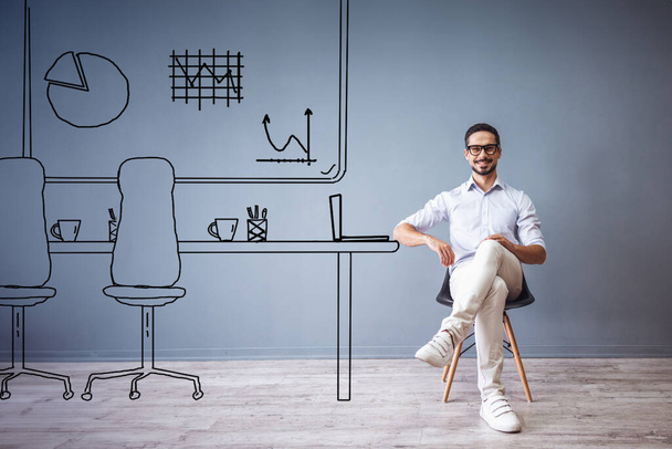 Knappe man in slimme casual kleding werkt met laptop terwijl zitten op de stoel op grijze achtergrond met getrokken kantoor - Foto, afbeelding