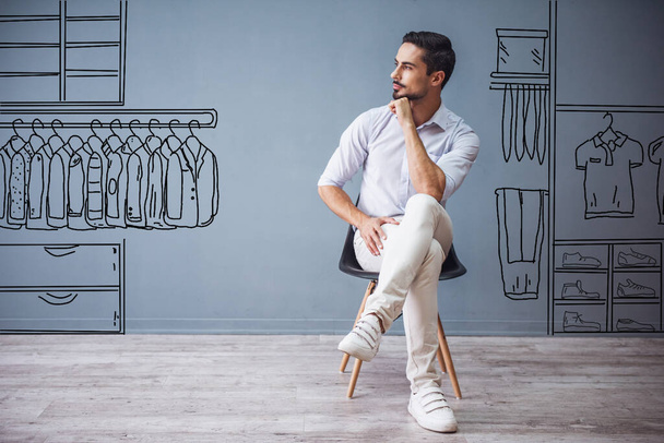 Όμορφος άντρας με έξυπνα casual ρούχα κοιτάζει αλλού και σκέφτεται ενώ κάθεται στην καρέκλα σε γκρι φόντο με ζωγραφισμένο κατάστημα ανδρών - Φωτογραφία, εικόνα