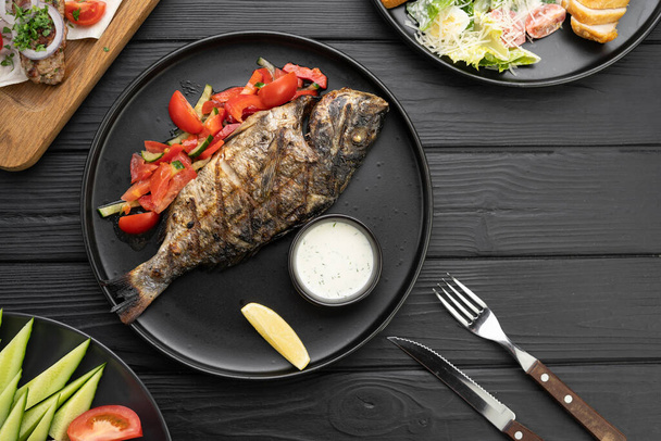 Restaurant menu concept. Delicious roasted dorado fish with lemon on black plate on dark wooden table, top view. Flat lay. Copy space - Photo, Image