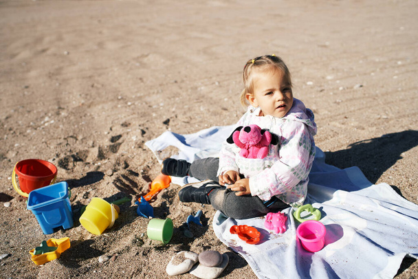 Pieni tyttö, jolla on leluja, istuu huopalla rannalla. Laadukas kuva - Valokuva, kuva