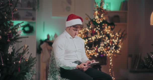 Junger Mann in Formalitäten und Weihnachtsmütze mit digitalem Tablet, während er zu Weihnachten im beleuchteten Haus auf einem Stuhl sitzt - Filmmaterial, Video