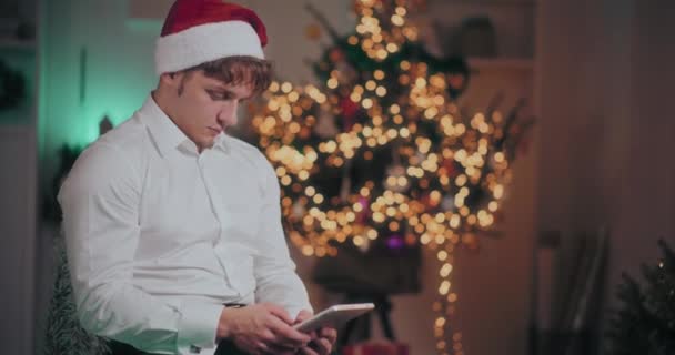 Fiatal férfi formális és Mikulás kalap segítségével digitális tabletta, miközben ül a megvilágított otthon karácsonykor - Felvétel, videó