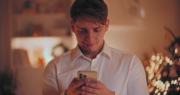 Hombre joven guapo mensajes de texto en el teléfono inteligente en casa durante la Navidad - Metraje, vídeo