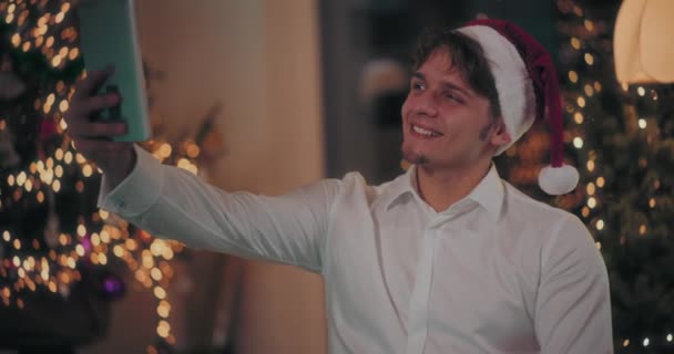 Ευτυχισμένος νεαρός άνδρας σε Σάντα καπέλο λαμβάνοντας selfie μέσω ψηφιακής tablet στο διακοσμημένο σπίτι κατά τη διάρκεια των Χριστουγέννων - Πλάνα, βίντεο