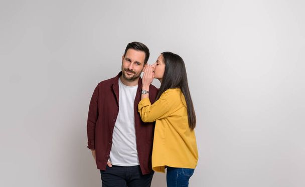 Joven novia susurrando secreto en la oreja guapo novio mientras que de pie sobre fondo blanco - Foto, Imagen