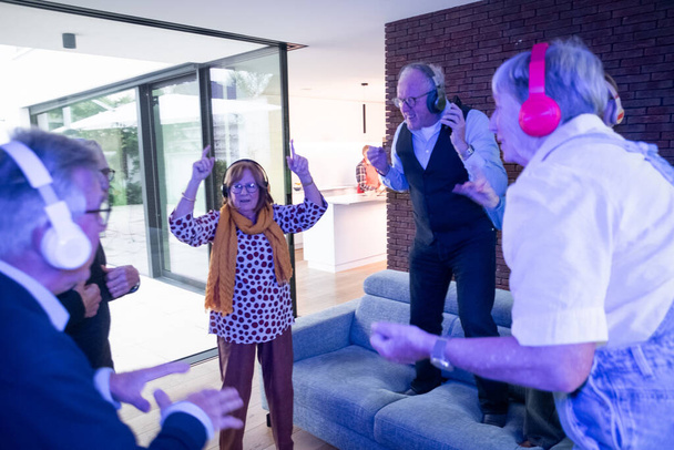 Een groep energieke senioren wordt gevangen midden in de dans, elk met een koptelefoon, waarschijnlijk genietend van een stille disco. De moderne woonkamer met zijn strakke lijnen en glazen deuren biedt een eigentijdse - Foto, afbeelding