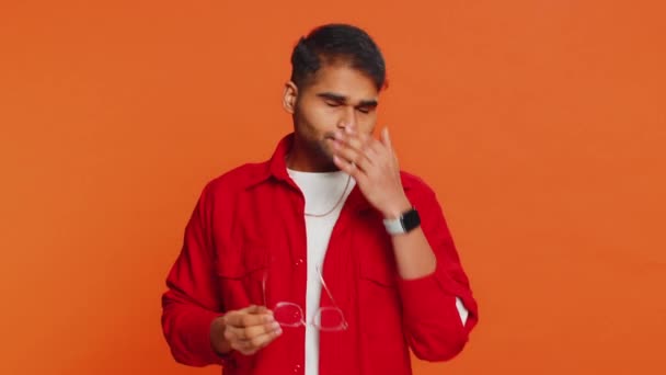 Exhausted tired Indian young man takes off glasses, feels eyes pain, being overwork burnout from long hours working. Sleepy exhausted Arabian guy rubbing eyes isolated on studio orange background - Footage, Video