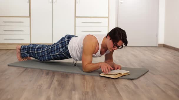 Jonge atletische man staat in de elleboog plank, trainen in de ochtend in de huiskamer.en het lezen van een boek Hoge kwaliteit 4k beeldmateriaal - Video