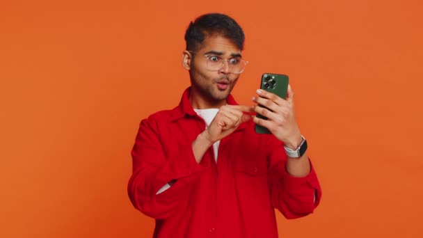 Glücklich aufgeregt indischen jungen Mann Kerl mit Smartphone tippen Browsing schreien sagen wow ja herausgefunden großen großen Gewinn gute Nachrichten Lotterie Ziel Erfolg feiern, gewinnen Spiel auf orange - Filmmaterial, Video