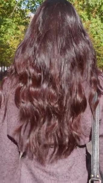 Mladá krásná brunetka s kudrnatými zdravými dlouhými vlasy a kabelkou se prochází v parku. Zpomal. Zadní pohled. Svislé video - Záběry, video