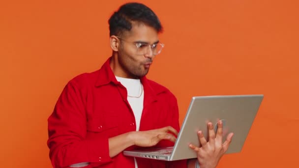 Glücklich aufgeregter indischer Mann tippt am Laptop, arbeitet an einem Projekt, genießt Ergebnisse, gewinnt Lotteriespiel, feiert Erfolg, Online-Shopping, Sieg, gute E-Mail-Nachrichten. Arabischer Kerl auf orangefarbenem Hintergrund - Filmmaterial, Video
