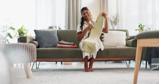 Frau auf Sofa zu Hause mit sauberer Wäsche, Hausarbeit und Wochenendarbeiten im Wohnzimmer mit Lächeln. Wartung, Reinigung und glückliches Mädchen sitzt morgens auf der Couch und faltet frische Kleidung in der Wohnung - Filmmaterial, Video