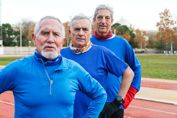 Trois hommes seniors en tenue de sport se tiennent fièrement sur une piste de course, respirant la confiance et la camaraderie après une séance d'entraînement. - Photo, image