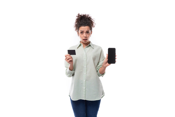 mujer rizada de pelo negro bastante joven con estilo de moño casual en camisa que sostiene maqueta de tarjeta de crédito de plástico y teléfono inteligente. - Foto, Imagen