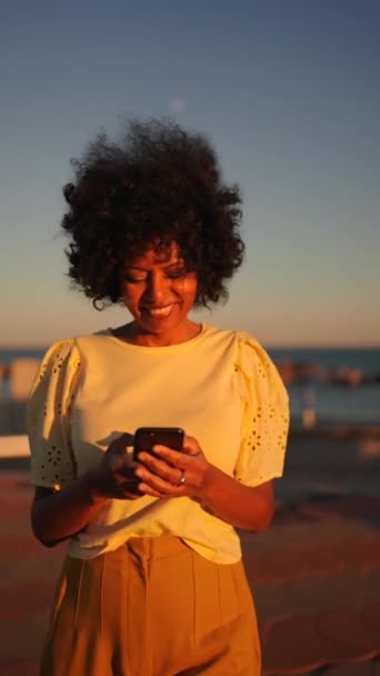 Video de una mujer sonriente usando el teléfono junto al mar al atardecer - Metraje, vídeo