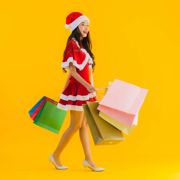 Портрет красива молода азіатка носить різдвяний капелюх з великою кількістю сумки для покупок на жовтому ізольованому фоні
 - Фото, зображення