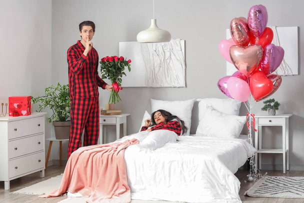 Młody mężczyzna z różami pokazując gest milczenia i jego śpiąca dziewczyna w sypialni w Walentynki - Zdjęcie, obraz