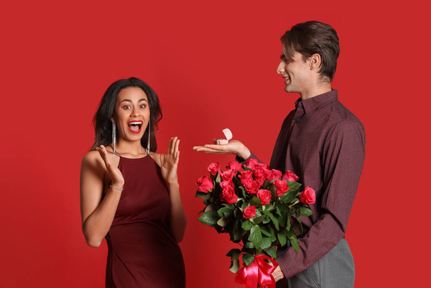 Νεαρός με τριαντάφυλλα και δαχτυλίδι που κάνει πρόταση γάμου στην κοπέλα του σε κόκκινο φόντο. Γιορτή του Αγίου Βαλεντίνου - Φωτογραφία, εικόνα