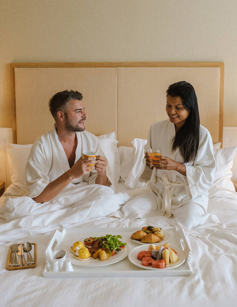 豪華なホテルでベッドで朝食をとるカップル, 朝食コーヒーとパンのベッドで男女. ベッドルームにいるアジア人女性とヨーロッパ人男性の - 写真・画像
