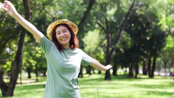 Retrato joven mujer asiática con sombrero de la mañana soleado que es alegre alegre feliz en un buen estado de ánimo asertivo positivo saltando en un jardín con árboles sombreados naturaleza aire puro sonriendo mirando a la cámara. - Metraje, vídeo