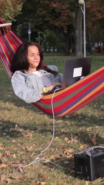 Nuori naisohjelmoija työskentelee kannettavan tietokoneen kanssa puun alla ja lataa sitä kannettavasta latausasemasta. Pystysuora video - Materiaali, video