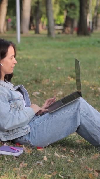 Mujer joven con un portátil sentado en la hierba en el parque en un día de otoño. Educación en reposo, tecnología y concepto de internet. Vídeo vertical - Imágenes, Vídeo