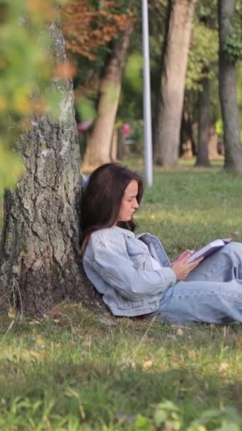 若い学生の少女が公園の草の上にコンピュータで座っている. 大学で勉強する. 教育とリモートワーキングコンセプト。 バーティカルビデオ - 映像、動画