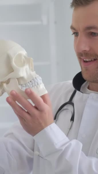 Szalenie wesoły młody, atrakcyjny lekarz trzymający w rękach ludzki szkielet czaszki. Szczęśliwy, pozytywny lekarz w nowoczesnej klinice ortopedycznej. Pionowe wideo - Materiał filmowy, wideo