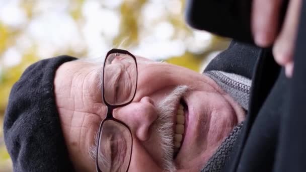 Ένας γέρος κάθεται σε ένα παγκάκι, κοιτάζοντας το κινητό του και γελώντας. Χρησιμοποιώντας gadgets σε ηλικία συνταξιοδότησης. SMS κειμένου, κινητή βιντεοκλήση, συνομιλία. Κάθετη βίντεο - Πλάνα, βίντεο
