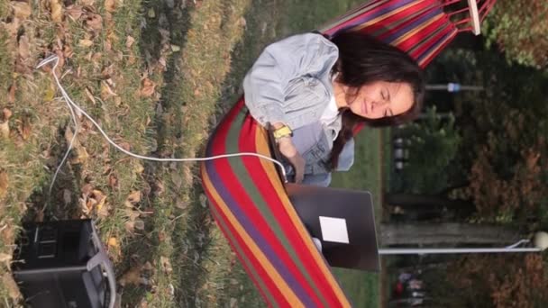 Genç bir kadın programcı bir ağacın altında dizüstü bilgisayar üzerinde çalışıyor ve onu taşınabilir şarj istasyonundan şarj ediyor. Dikey video - Video, Çekim