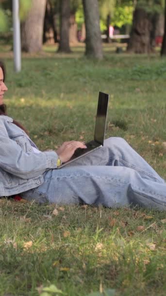 Nuori kaunis tyttö kannettavan kanssa lepäämässä puistossa. Nuori nainen työskentelee tietokoneella puistossa istuessaan peiton päällä. Pystysuora video - Materiaali, video
