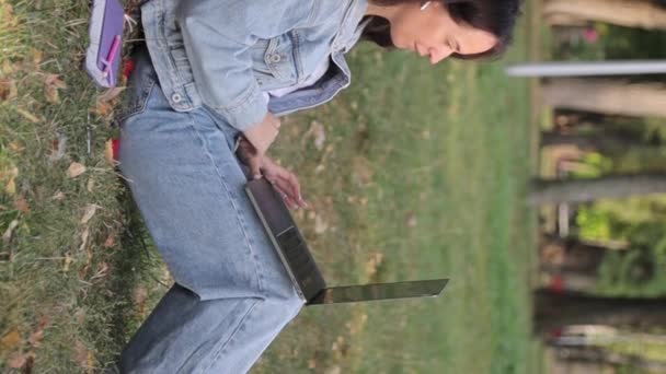 Młoda kobieta z laptopem siedząca na trawie w parku w jesienny dzień. Odpoczynek edukacji, technologii i koncepcji Internetu. Pionowe wideo - Materiał filmowy, wideo