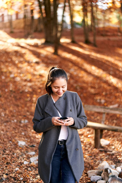 Улыбающаяся девушка смотрит в кружку с чаем в руках в осеннем парке. Высокое качество фото - Фото, изображение